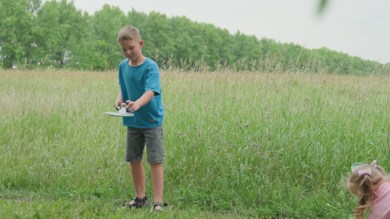 흰색 셔츠를 입은 소년이 야외에서 배드민턴을 치고 있어요. 파란색 셔츠를 입은 십 대가 키 큰 풀밭 위로 셔틀콕을 치고, 집중된 자세로 운동감 넘치는 스윙을 하고 있어요. 부드러운 햇빛 아래 초원에서 혼자 훈련하며 활기찬 움직임을 보여줍니다.