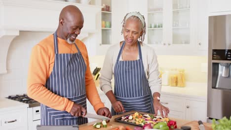 african american senior couple in aprons preparing meal using tablet in kitchen, slow motion