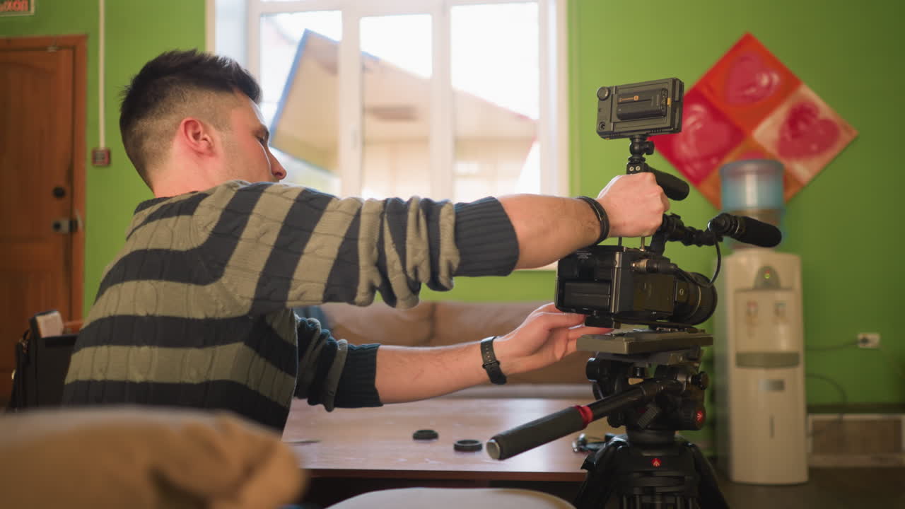 Back view of Studio guy adjusting camera on tripod with microphone attached, preparing for video shoot in home studio. Camera gear and accessories laid out on table for filming project
