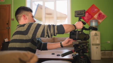 back view of studio guy adjusting camera on tripod with microphone attached, preparing for video shoot in home studio. camera gear and accessories laid out on table for filming project