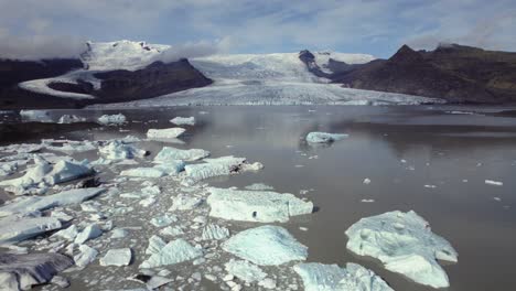 Aerial-flying-over-dramatic-icebergs-floating-in-water,-Jokulsarlon-lake,-winter-snow-landscape,-glacier-in-distance,-Iceland