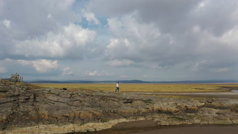 Toma-Aérea-Amplia-De-Una-Persona-Caminando-A-Lo-Largo-De-Las-Rocas-En-Una-Línea-De-Costa-Con-Marea-Baja,-En-Un-Brillante-Día-De-Verano