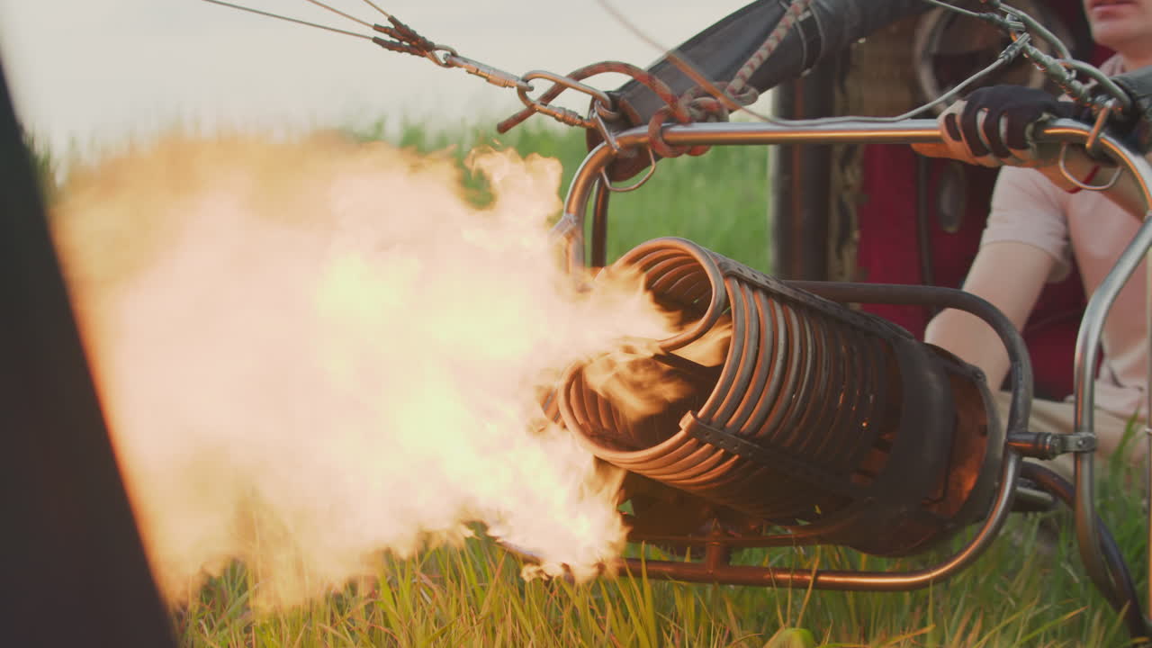 Partial view of man operating burner releasing strong flame into balloon envelope during hot air balloon inflation on grassy field, showing coiled metal structure, and intense heat energy