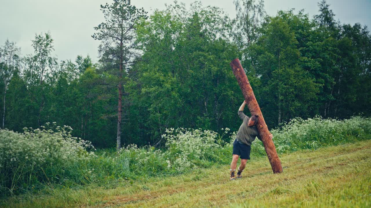 A Man Pushes a Tall Wooden Log Upright in a Grassy Field Near the Forest - Static Shot