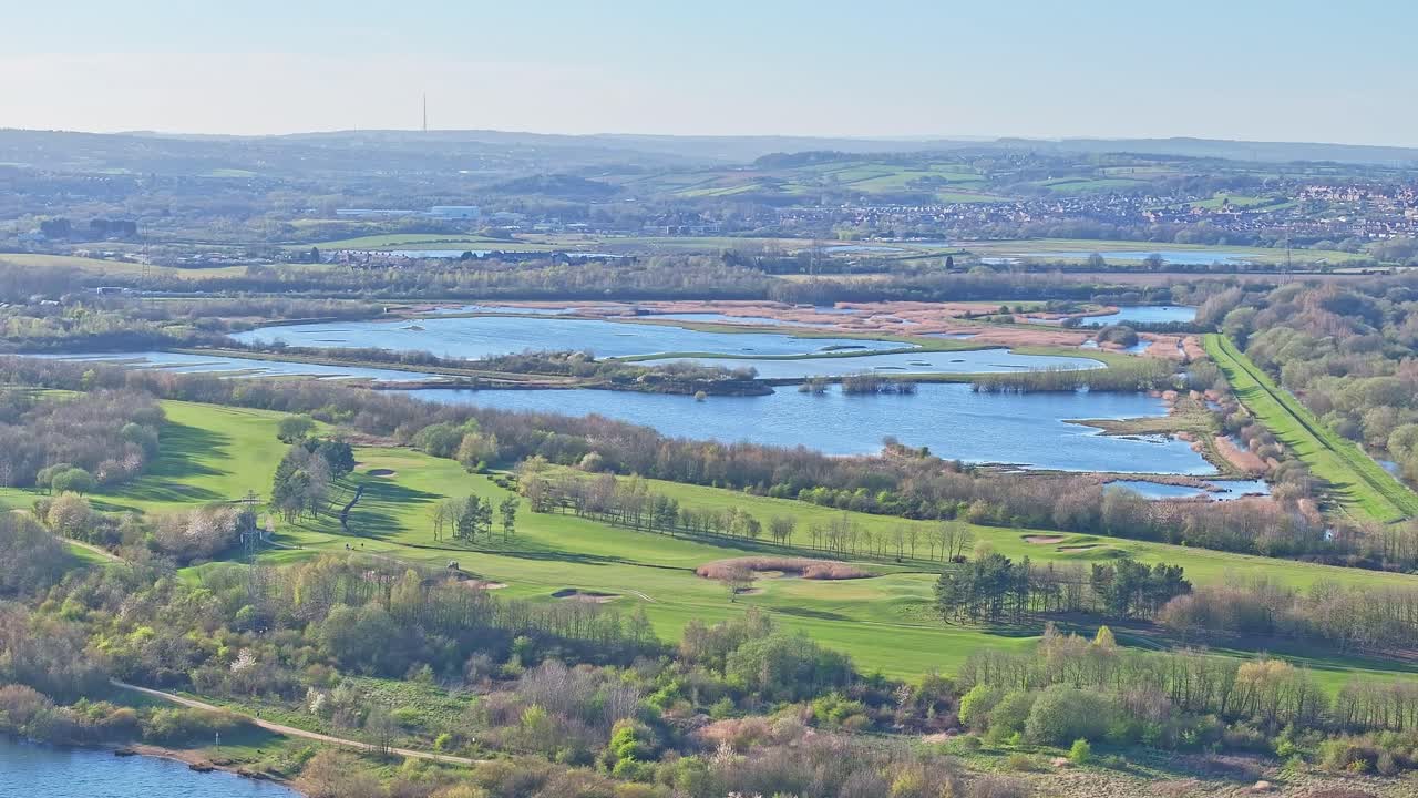 Drone captures Manvers Lake, adjacent golf course and wetland reserve, with lush green fairways, water channels, reeds, and hilly South Yorkshire countryside under morning light, real time