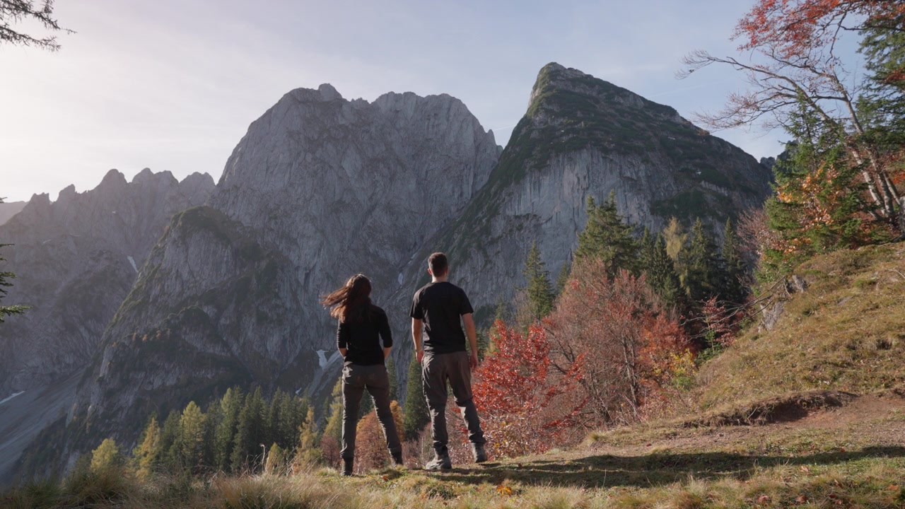 Due individui stanno insieme, ammirando la vista mozzafiato di montagne aspre e alberi colorati mentre il sole tramonta in un sereno paesaggio autunnale