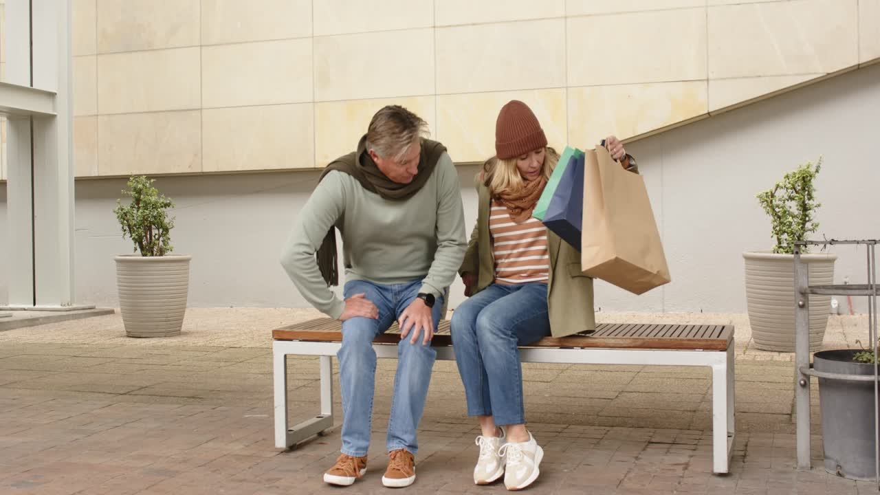 Senior couple walking on plaza toward bench carrying bags then sitting as man bending in pain