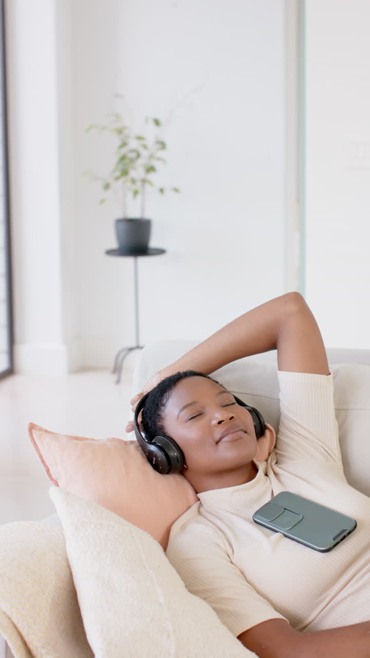 Vertical video of african american woman using headphones and smartphone on sofa, slow motion