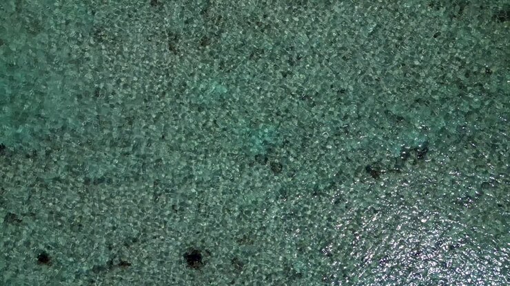 Drone footage flys backwards whilst slowly rising up over the rippling surface of a tropical blue ocean above a coral reef in the Caribbean
