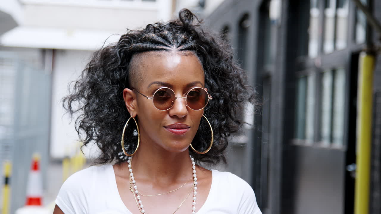 Giovane donna afroamericana alla moda che si gira e guarda la fotocamera sopra i suoi occhiali da sole rotondi, ridendo, primo piano