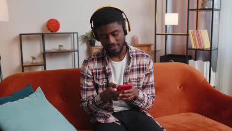 Overjoyed-young-man-in-wireless-headphones-dancing,-singing-on-cozy-couch-in-living-room-at-home