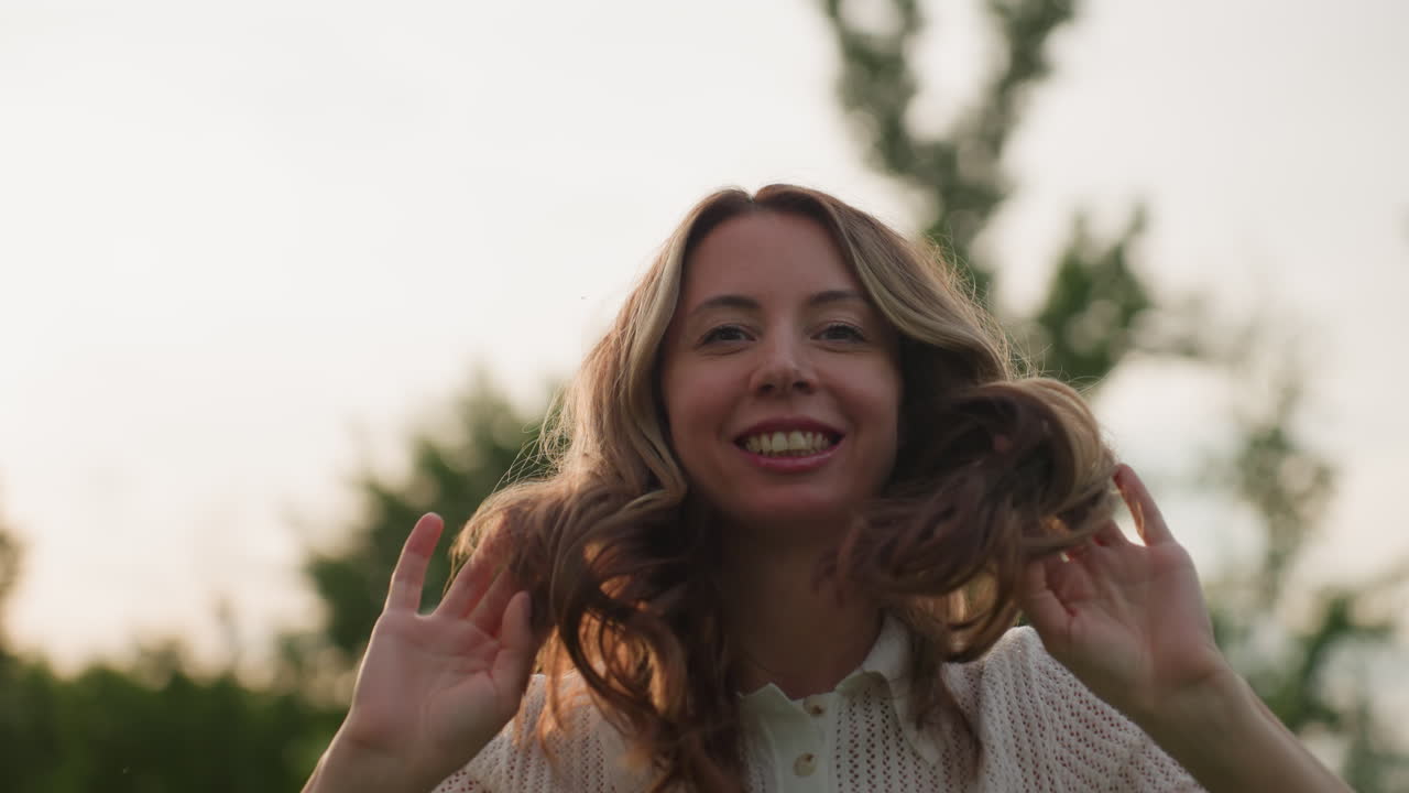 portrait of beautiful woman with smiles posing with hands in hair, soft backlight from sky, blurred tree background, confident calm gaze, subtle smile, outdoor summer setting showing elegance