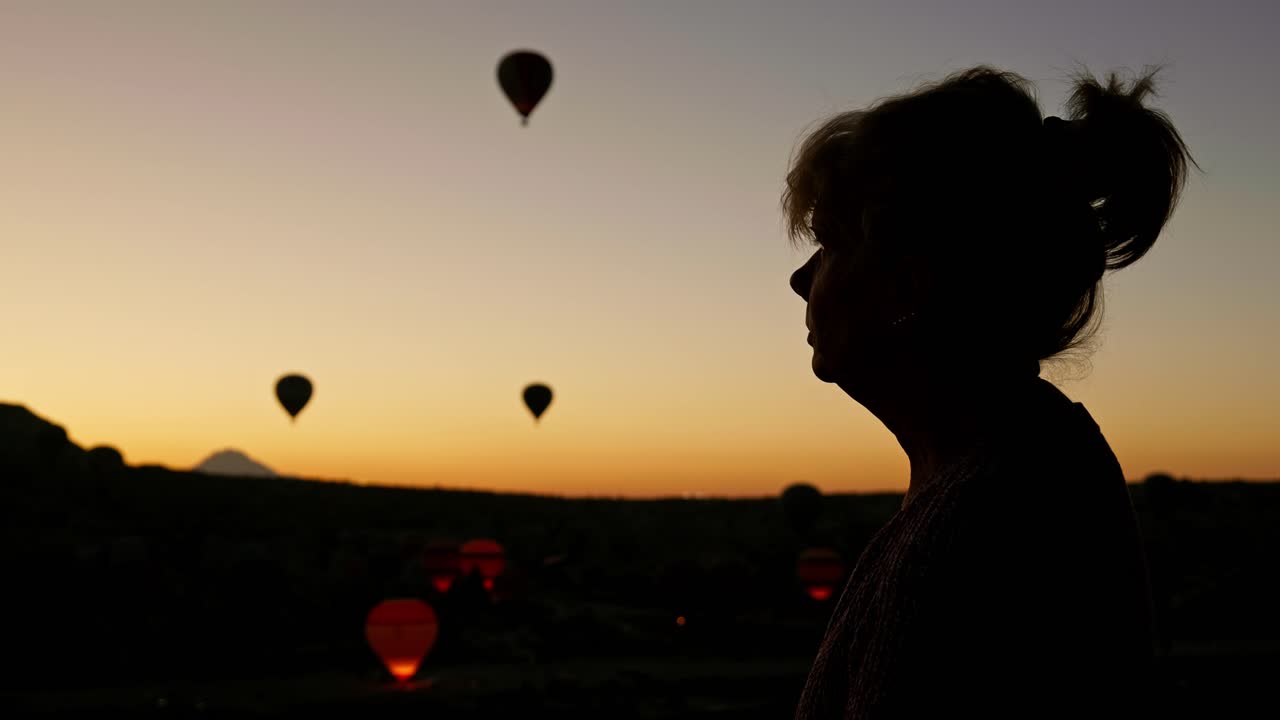 Mulher em silhueta observa balões de ar quente ao nascer do sol de manhã cedo.