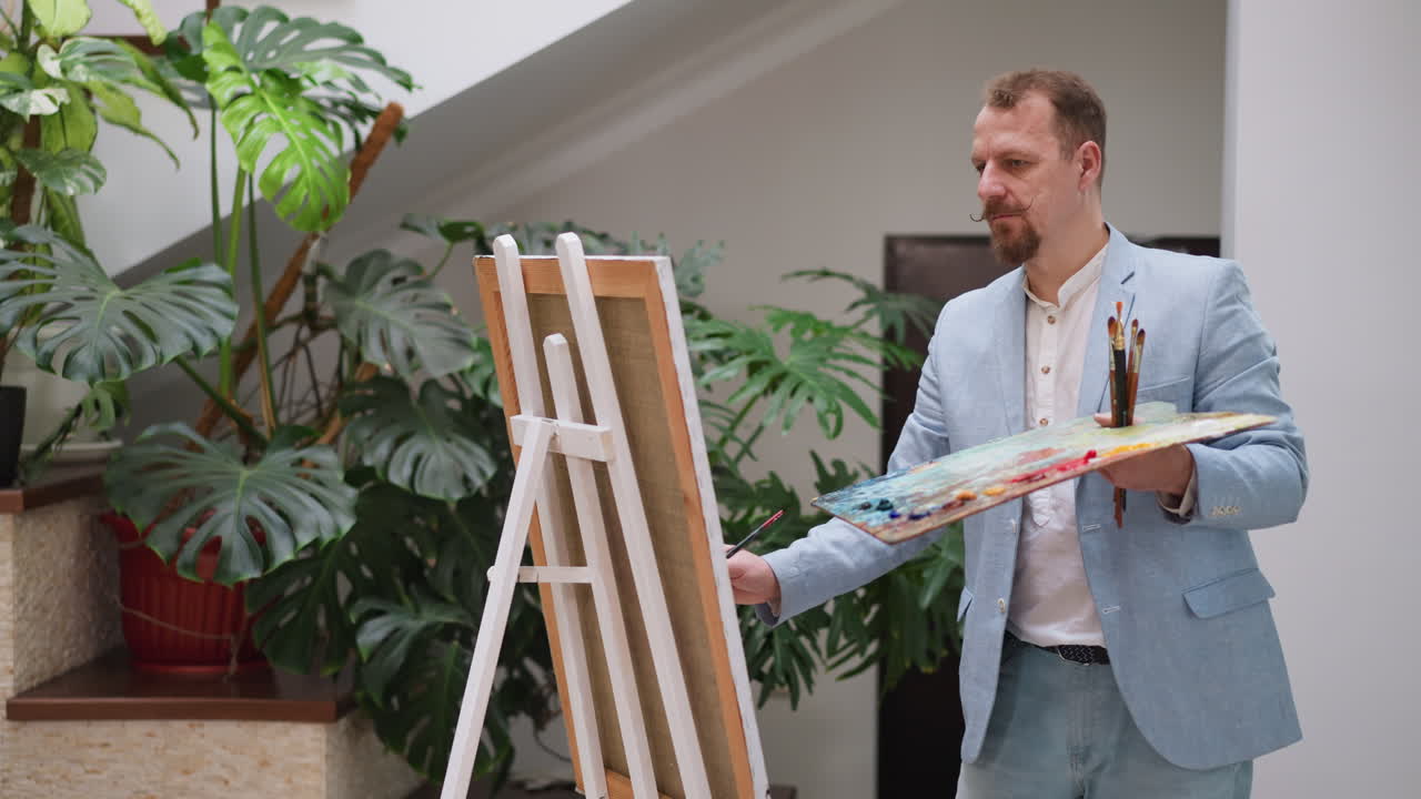 Art master draws on canvas in studio. Stylish artist in blue stylish suit creates picture holding palette by easel. Mature painter enjoys creativity in shop