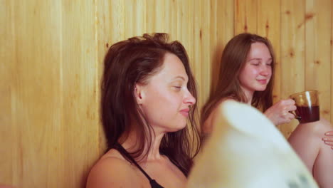 close up of young woman fanning herself with sauna hat while two blurred women in background enjoy steamy atmosphere, chatting and toasting drinks in warm wooden interior filled with relaxing energy