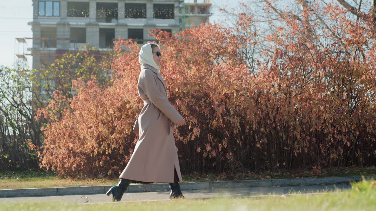 Donna solitaria con foulard che cammina in un parco autunnale, lungo soprabito rosa, piccolo albero sempreverde, arbusti arancioni, cantiere edile lontano, passo lento e riflessivo, atmosfera diurna fresca, profilo laterale, delicata