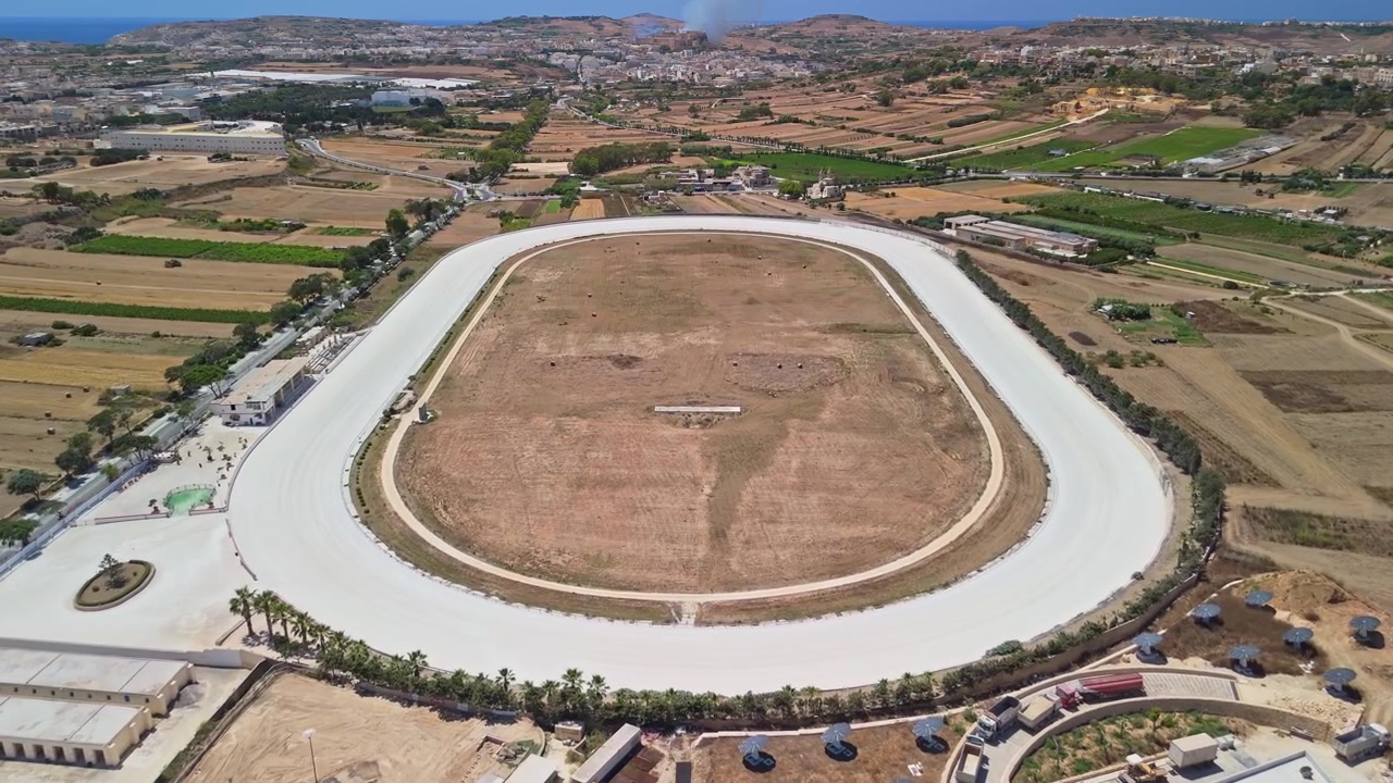 Xewkija, Gozo horse racing track and sports venue surrounded by farmland, roads and buildings on the Maltese island landscape, captured by drone in sunny summer weather.