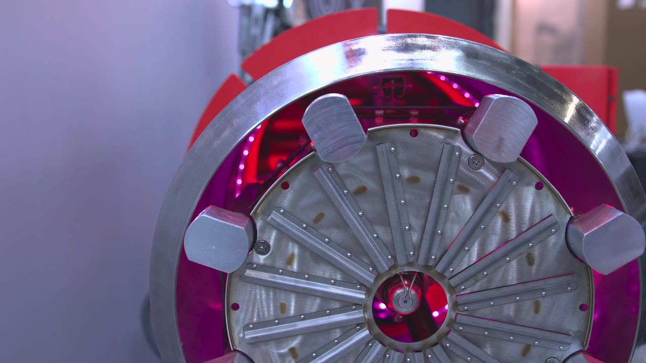Un primo piano frontale mostra un rotore metallico circolare fissato da molteplici ganasce di serraggio esagonali all'interno di un alloggiamento verniciato di rosso, con apertura interna esposta, cablaggio, bulloni e superfici spazzolate riflettenti illuminate da una luce vivida