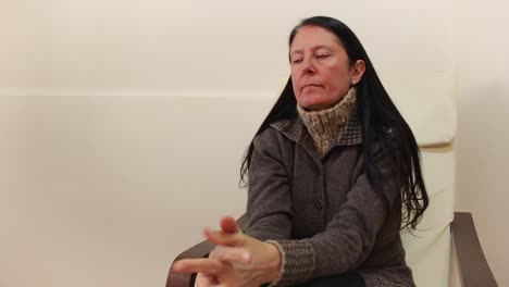 middle-aged woman seated in armchair looking at her hands nervously