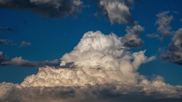 Time lapse of a towering white cumulus cloud rises above a layer of darker clouds against a deep blue sky, creating a dramatic atmospheric scene.
