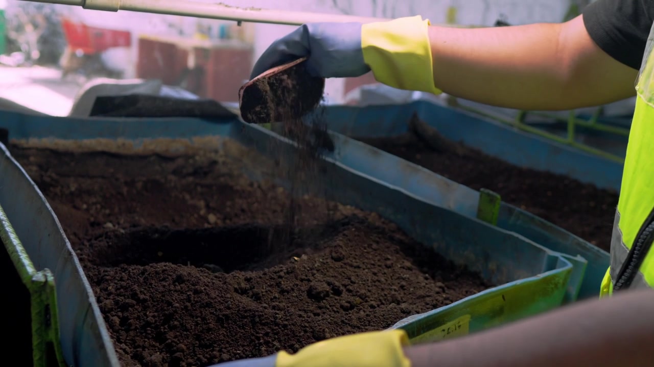 Close-up of a worker in gloves stirring dry black soil, preparing it for organic compost in a circular agribusiness project focused on sustainability and waste management.