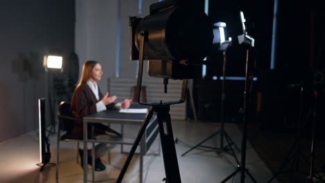 soffits illuminate female reporter talking to camera. woman in brown jacket sits at desk and speaks waving her hands.