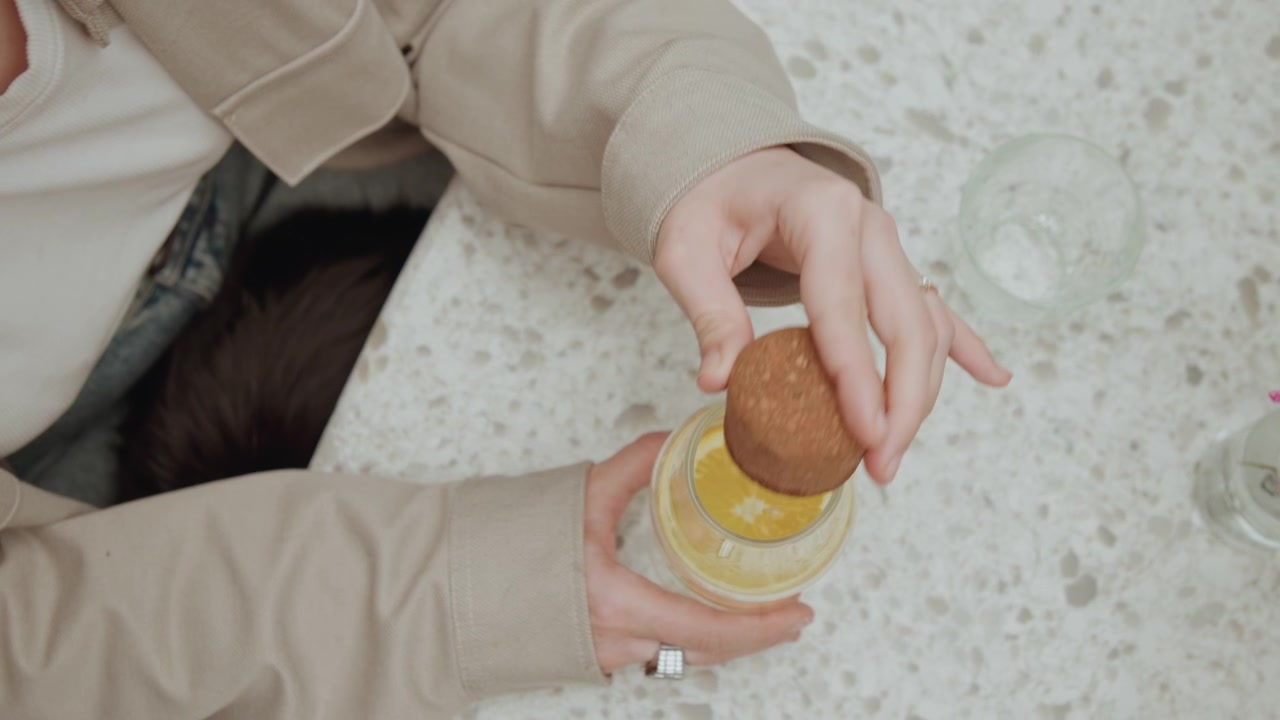 Primo piano di una donna con giacca beige che rimuove un tappo da un barattolo con liquido giallo e una fetta di limone. La scena include bicchieri vuoti, fiori rosa sul tavolo, illuminazione soffusa, espressione calma e dettagli della texture.