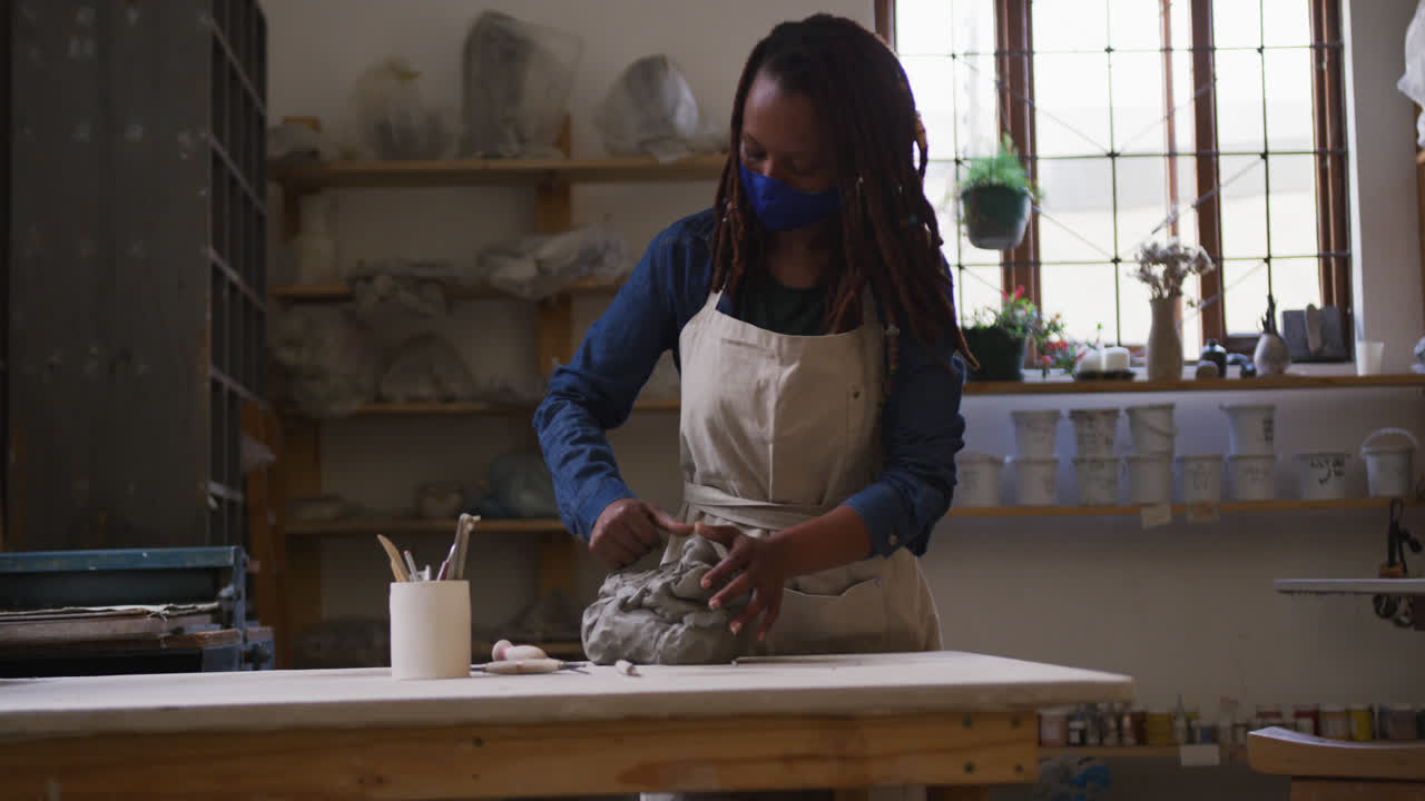 Female potter wearing face mask and apron kneading the clay at pottery studio