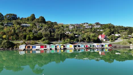 drone footage captures vibrant boathouses reflecting on calm waters in akaroa, new zealand, under clear blue skies