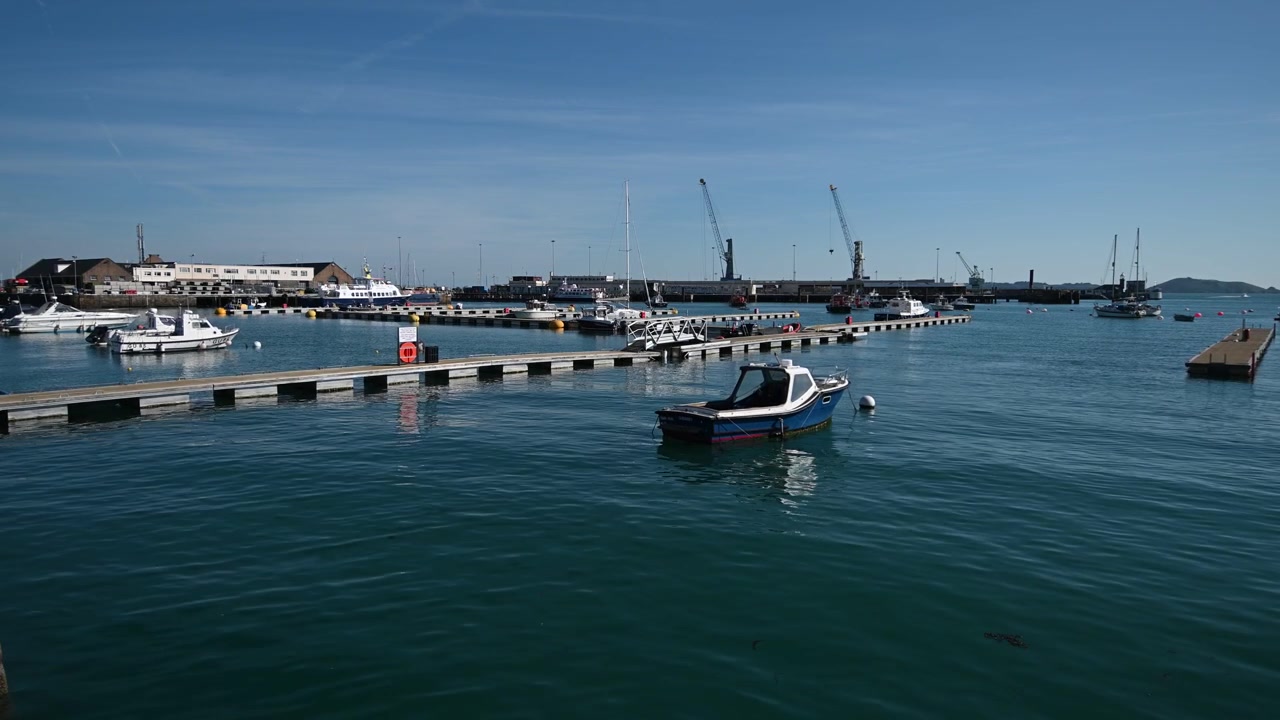 Hafen St. Peter Port, Kanalinseln Guernsey. Ansicht auf Wasserhöhe eines kleinen Bootes, das im Hafen schaukelt, mit Blick über Pontons auf Cambridge Berth und die Docks, wobei Jethou durch die Hafeneinfahrt sichtbar ist.