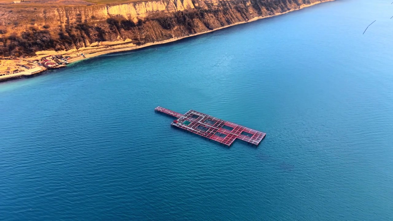 Floating aquaculture cage in calm Black Sea bay, Bulgaria. Overhead drone view, large rectangular red grid fish farm structure centered on turquoise water, limestone cliff and shore.