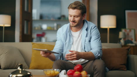 Carefree-gentleman-sitting-evening-home.-Positive-guy-taking-tablet-computer