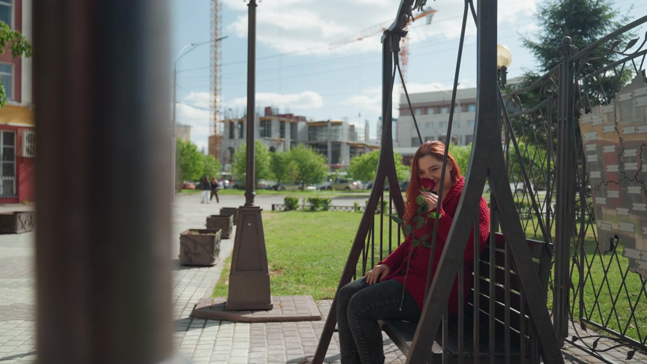 Donna con maglione rosso siede su un'altalena all'aperto, tenendo in mano e annusando foglie verdi. La scena presenta un ambiente urbano con edifici, alberi e gru edili sotto una luce diurna intensa. Ombre tenui.