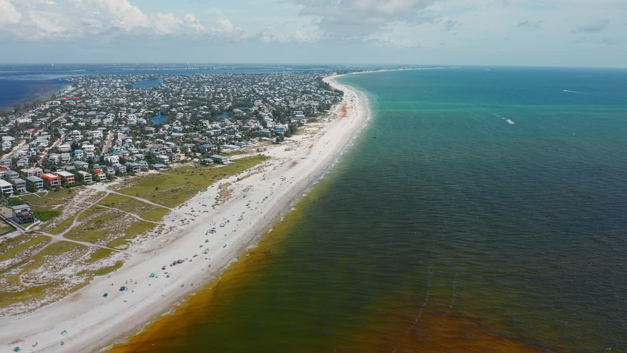 An aerial perspective of Anna Maria Island highlights its long sandy shoreline, vibrant turquoise waters blending with darker seas, and a coastal community stretching into the horizon
