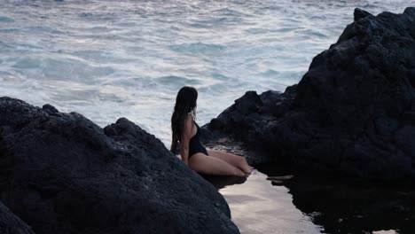 woman sits by volcanic rocks with her feet in a tide pool, gazing at the ocean waves in seixal, madeira.