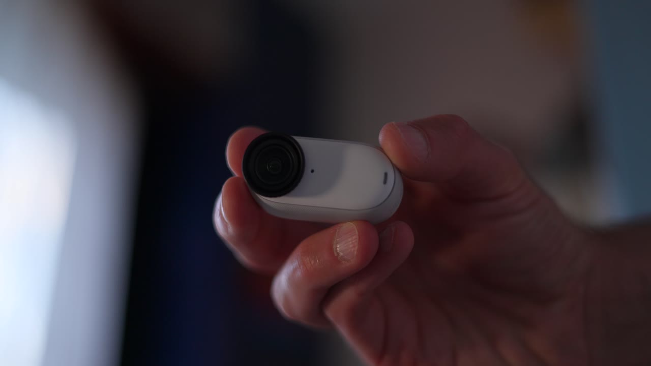 Hand rotating small, white, modern Insta360 GO 3S camera with a black lens and a red light indicator, demonstrating compact and innovative technology in everyday life