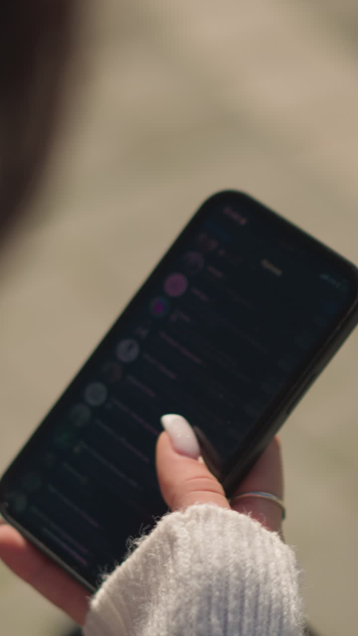primer plano de una mujer con cabello corto en abrigo negro y suéter blanco desplazándose por el teléfono con enfoque en la mano sosteniendo el teléfono con manicura visible, luz exterior suave y fondo borroso