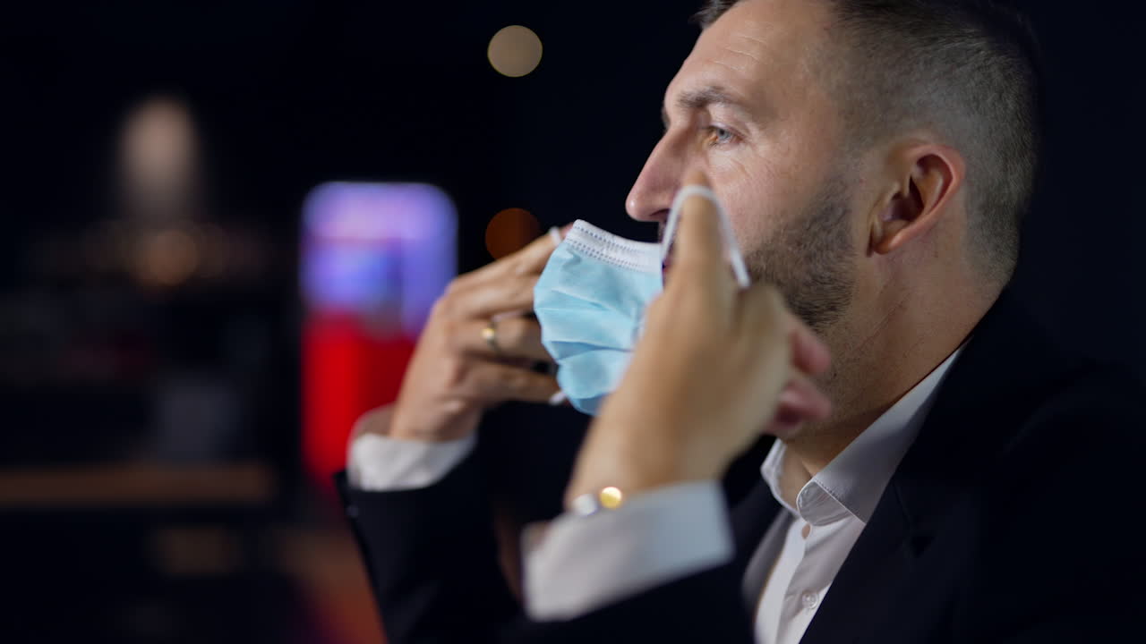 Bearded middle-aged man puts on surgical mask. Caucasian male side view portrait wearing mask during pandemic times. Dark blurred background.