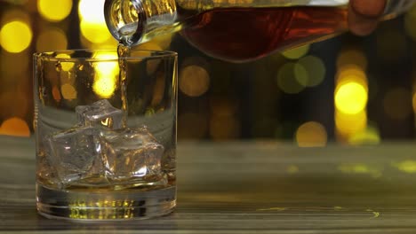 Gießen-Von-Goldenem-Whiskey,-Cognac-Oder-Brandy-Aus-Der-Flasche-In-Ein-Glas-Mit-Eiswürfeln.-Glänzender-Hintergrund