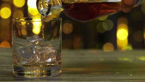 pouring of golden whiskey, cognac or brandy from bottle into glass with ice cubes. shiny background