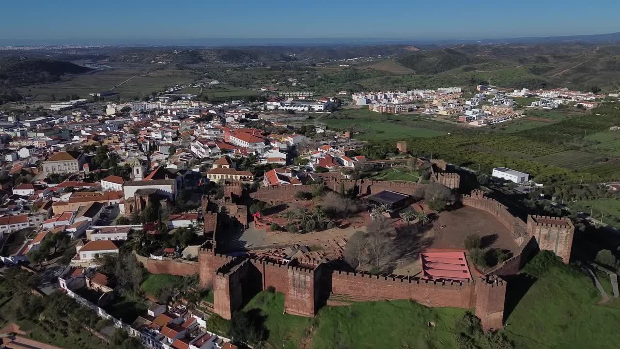 Drone view of historic red-brick castle overlooking rooftops and countryside in Faro, Portugal. Fort stands on green hilltop with panoramic view of nearby buildings, fields, and distant hills.