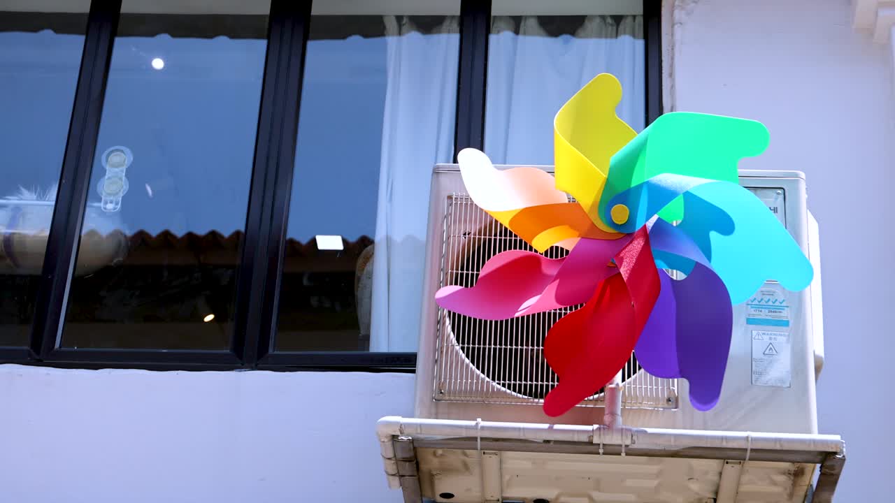 A vibrant plastic pinwheel rapidly spins atop an air conditioning unit outside a city window, captured in bright daylight with a static camera angle
