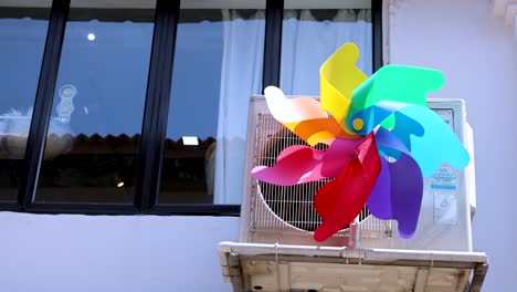 a vibrant plastic pinwheel rapidly spins atop an air conditioning unit outside a city window, captured in bright daylight with a static camera angle