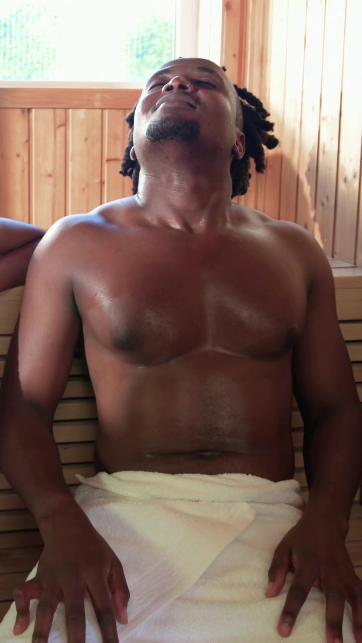 縦型動画：サウナの木製ベンチでタオルを巻いた男性がもたれかかり、熱気で体が揺れ動く様子。