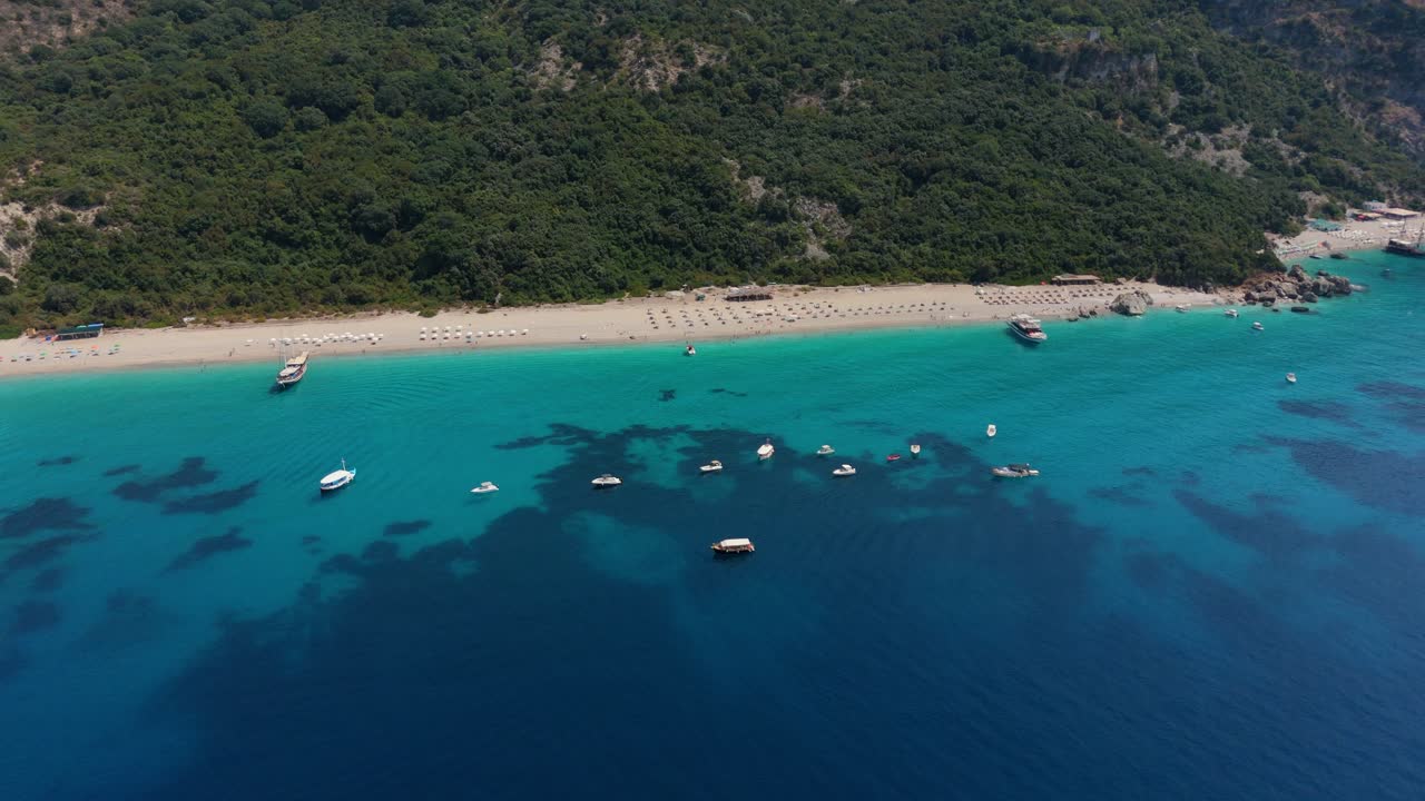 Turquoise waters and boats along a quiet sandy beach backed by lush green cliffs in Albania