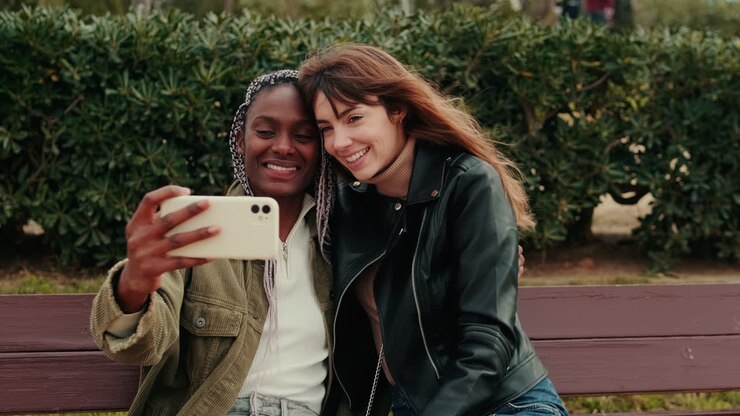 Due giovani donne felici che si scattano un selfie e si baciano su una panchina del parco