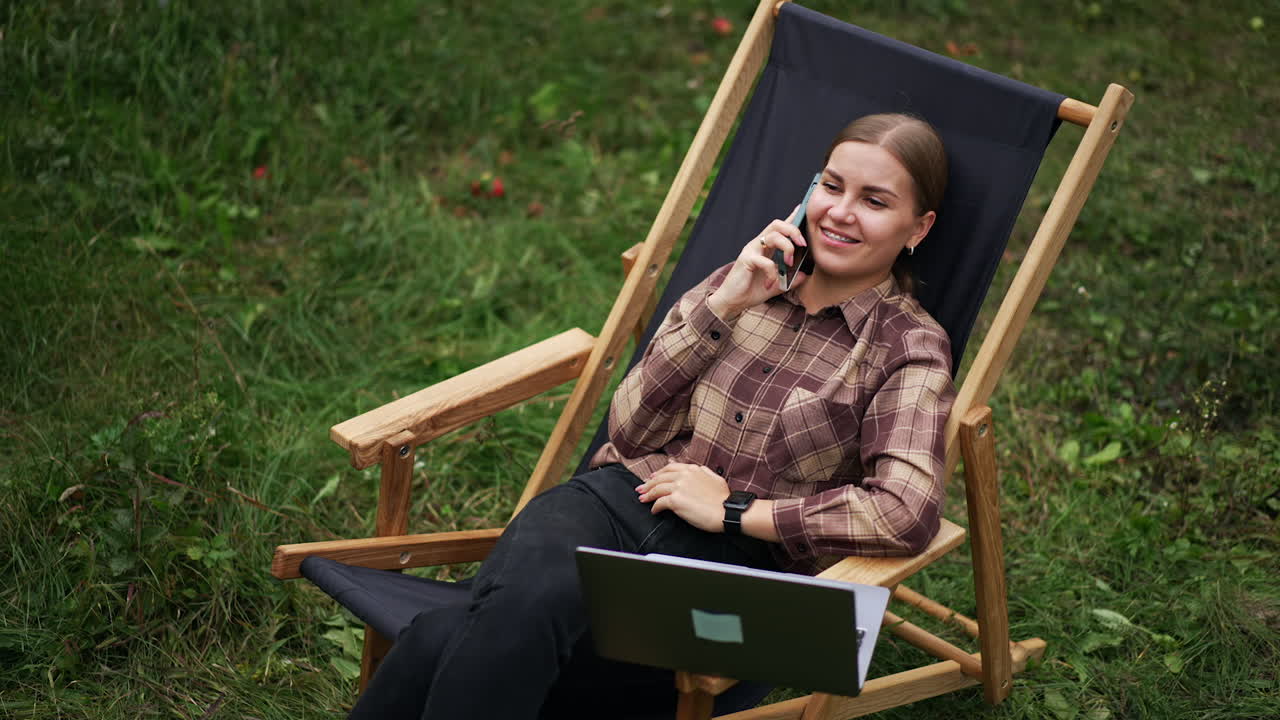 Caucasian relaxed lady speaks on the phone laying back in garden chair. Freelancing woman working remotely on laptop distracted from work to talk. Top view.
