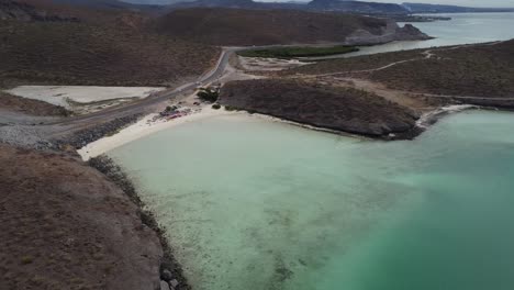 Aerial-panorama-view-in-front-of-the-beautiful-coast-of-Playa-El-Tecolote-in-Baja-California-Sur-Mexico-overlooking-the-calm-sea,-dry-landscape-and-a-road-right-on-the-coast-during-an-adventurous-trip