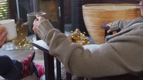 Mixed-race-couple-toasting-and-drinking-coffee-while-sitting-near-the-fireplace-at-vacation-home