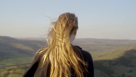 stabilised medium shot of young blonde woman standing in wind on top of mam tor, castleton, peak district, england before stopping to admire the view of green rolling hills and blue skies